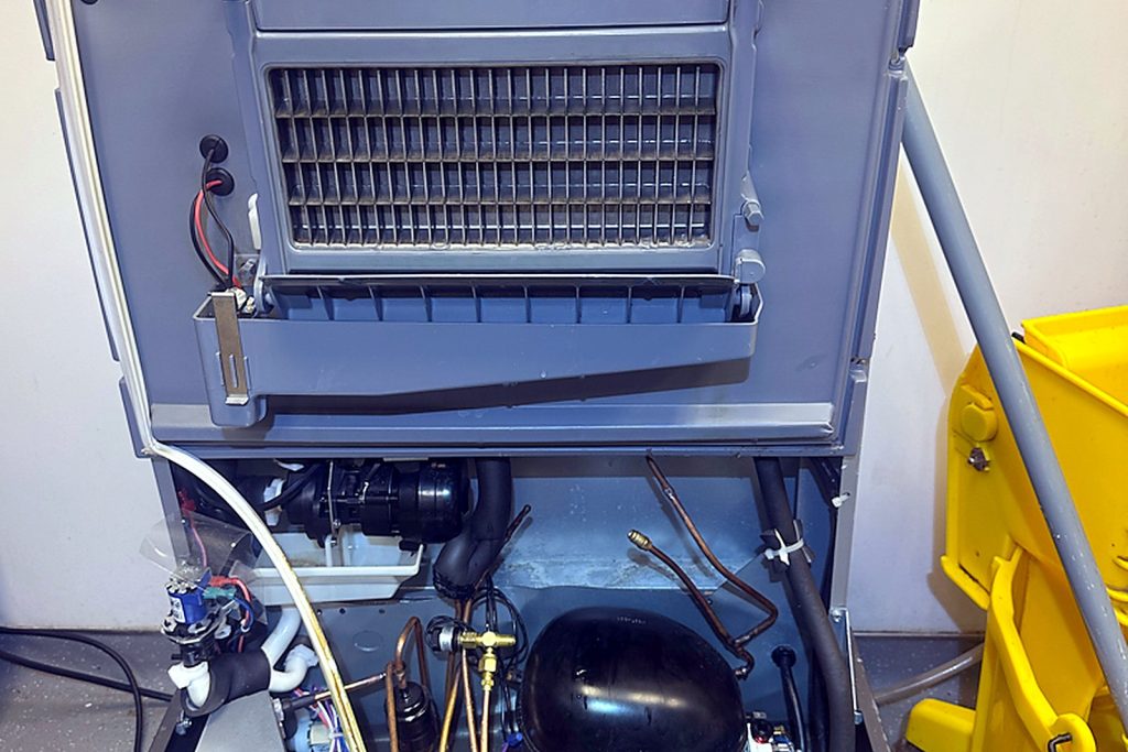Manitowoc UYF0140A undercounter ice machine pulled out and opened on a bakery floor, condenser coil and compressor exposed