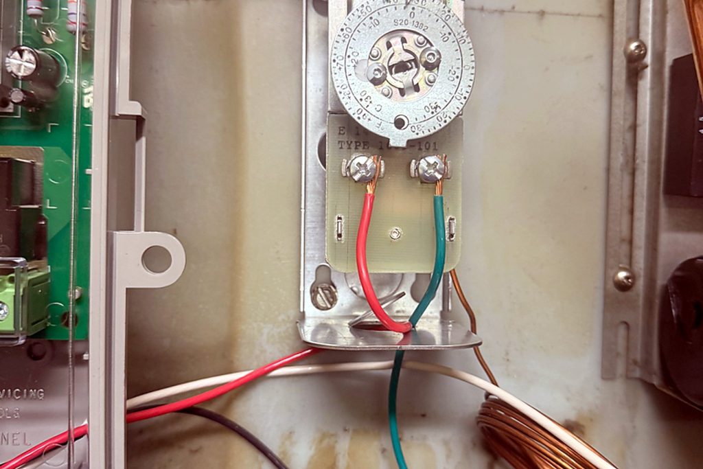 The original mechanical defrost timer, a clock-motor dial with trip pins, in the compressor compartment. The motor had stopped, so the dial never reached a defrost.