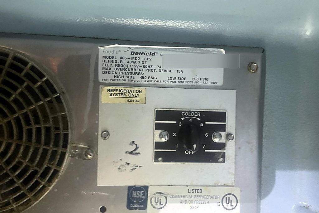 Delfield 406 series undercounter data plate listing R-404A and a 7 ounce charge beside the unit's dial thermostat, serial number redacted