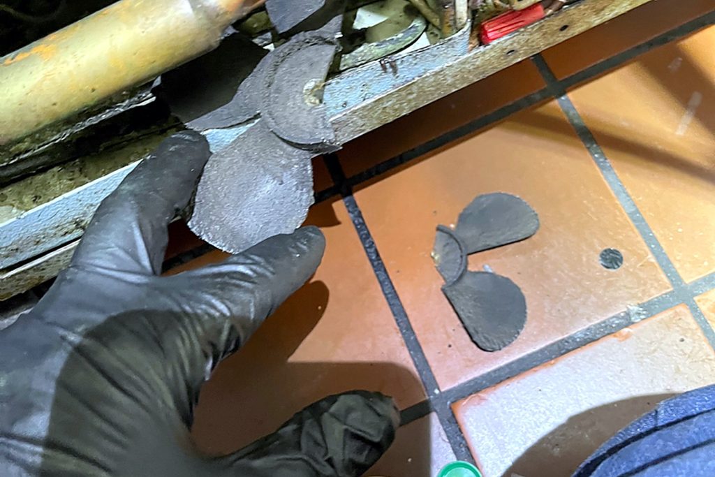 Gloved hand holding pieces of a shattered condenser fan blade, with more pieces on the floor beneath the unit