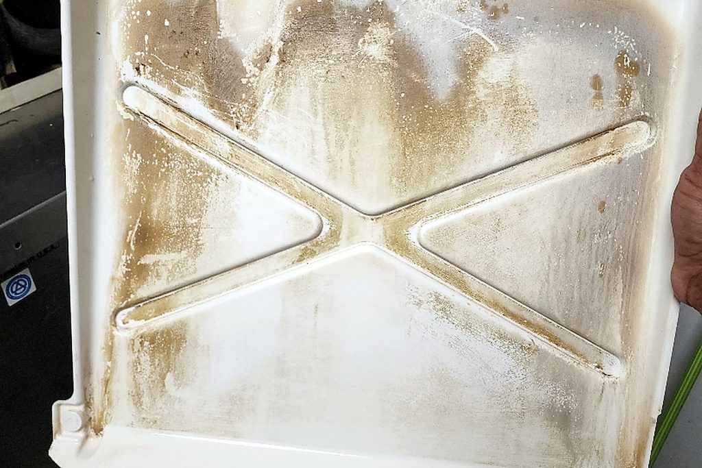 Ice machine water pan removed and held up, its surface coated in brown mineral scale