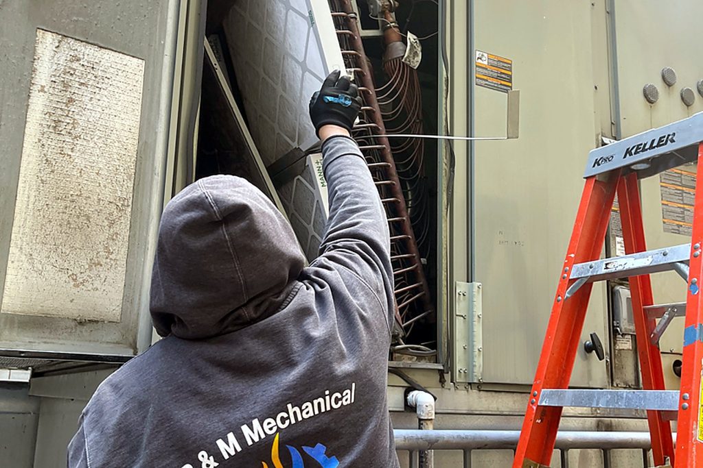 Technician in a B&M Mechanical hoodie on a ladder pulling a used pleated filter from the access panel of a rooftop unit